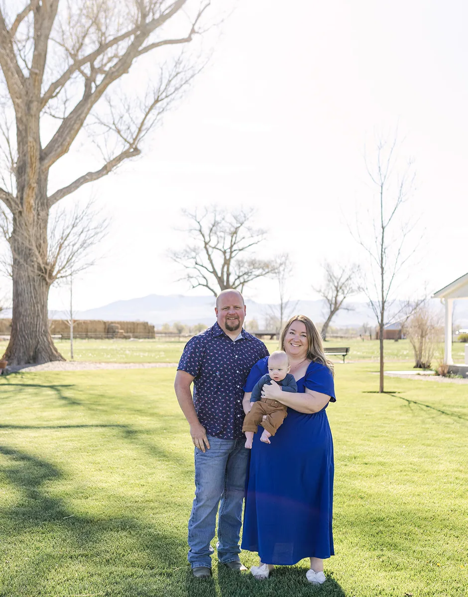 Kylie and Trevor with their baby, standing on a sunny lawn with trees and mountains behind them