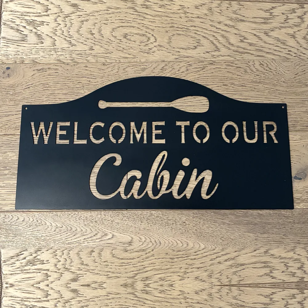 Black steel Welcome to Our Cabin sign with a canoe paddle.